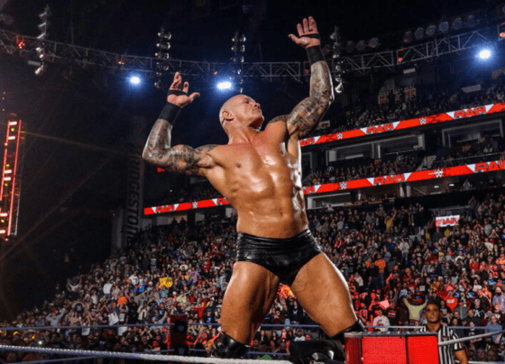 Randy Orton, a professional wrestler, striking a victorious pose on the turnbuckle in the wrestling ring, with the crowd cheering in the background.