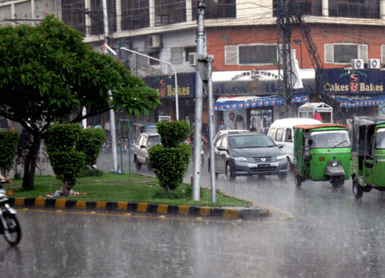 Heavy rainfall soaking a bustling street in Pakistan, with pedestrians using umbrellas and vehicles splashing through puddles.