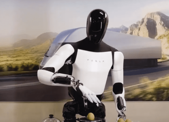 A life-size humanoid robot named Tesla Optimus, featuring a sleek, modern design with a primarily white and black color scheme. It stands upright, resembling a human in posture and proportions.